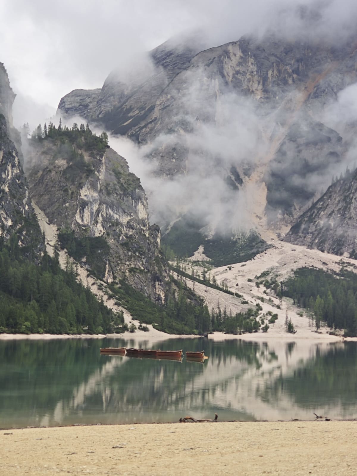 The shore of Lago di Braies, a row of wooden rowing boats pulled up at the far end under cloud-wreathed cliffs and a scree slope