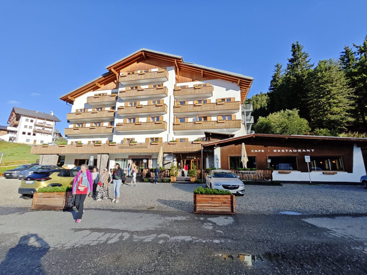 A South Tyrolean mountain hotel in morning sun — a white building with timber balconies and a restaurant terrace, pines behind it