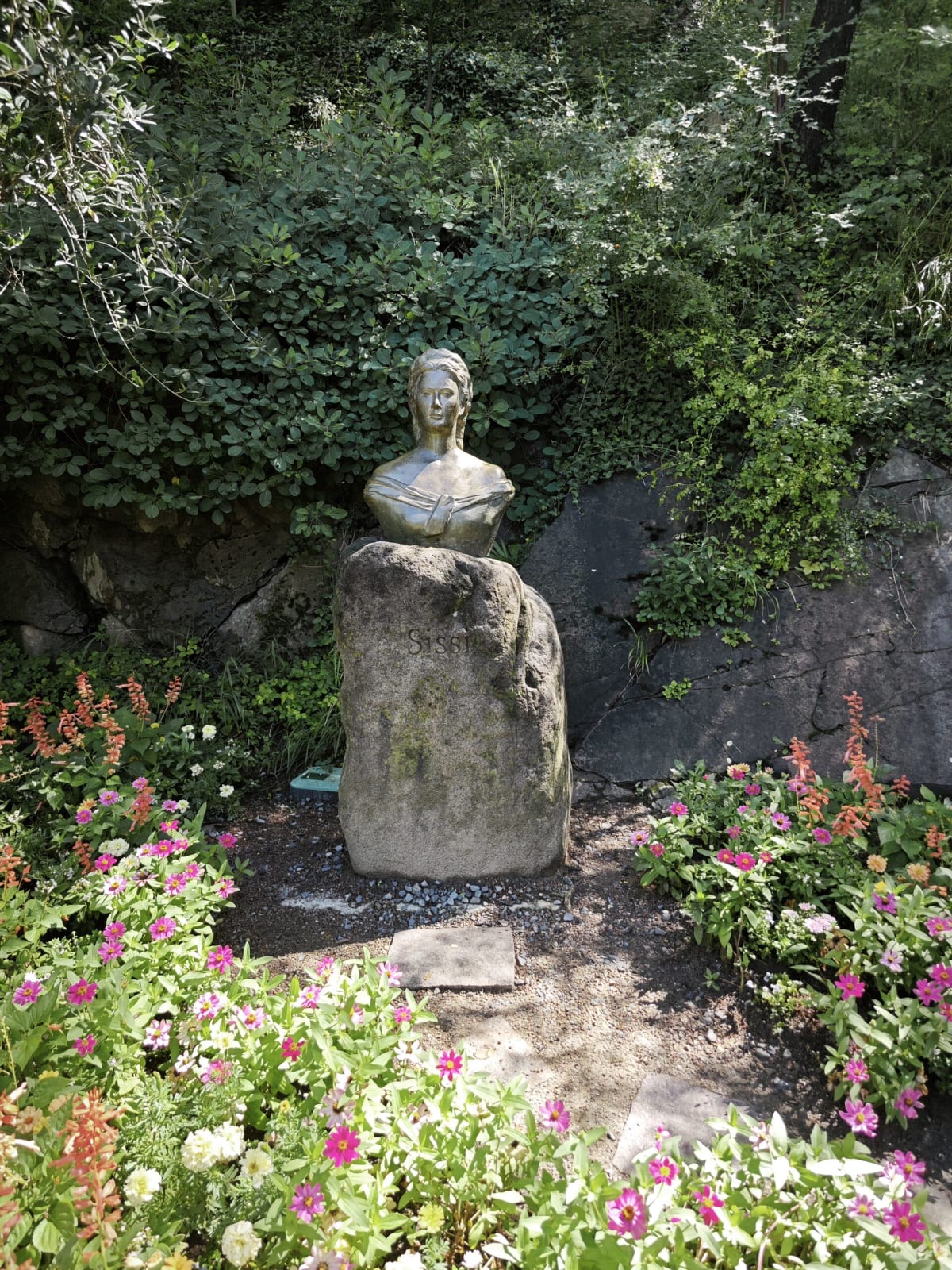 A stone bust on a rock among pink and orange flowers in a shady corner of the gardens