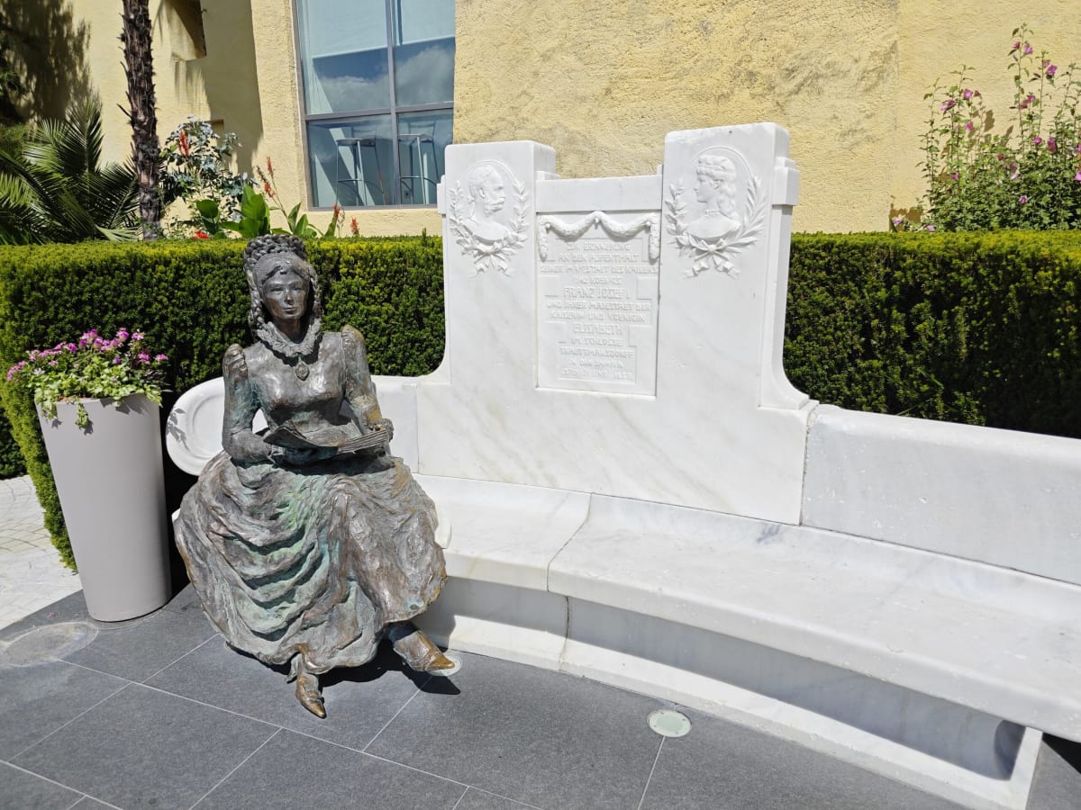 A bronze statue of a woman in a crinoline dress seated on a white marble bench, inscribed panels behind her and a yellow wall beyond