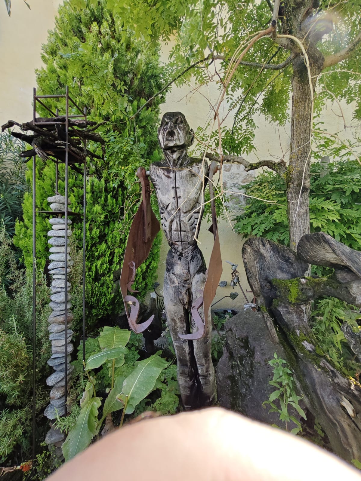 A metal sculpture of a gaunt figure hanging among plants and a chrome frame in the garden, a thumb at the edge of the frame
