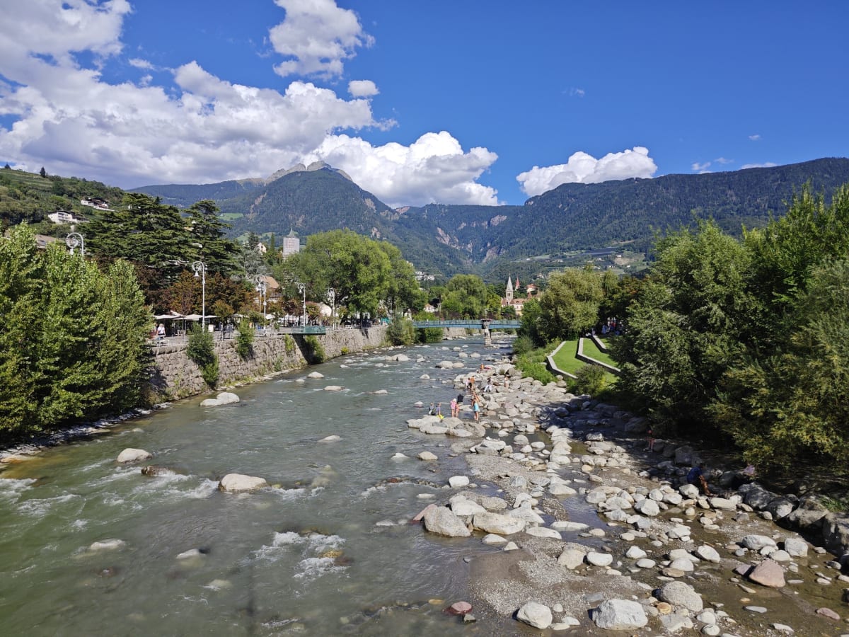 The Passer river tumbling over boulders through Merano, tree-lined banks and a promenade, mountains rising beyond the town