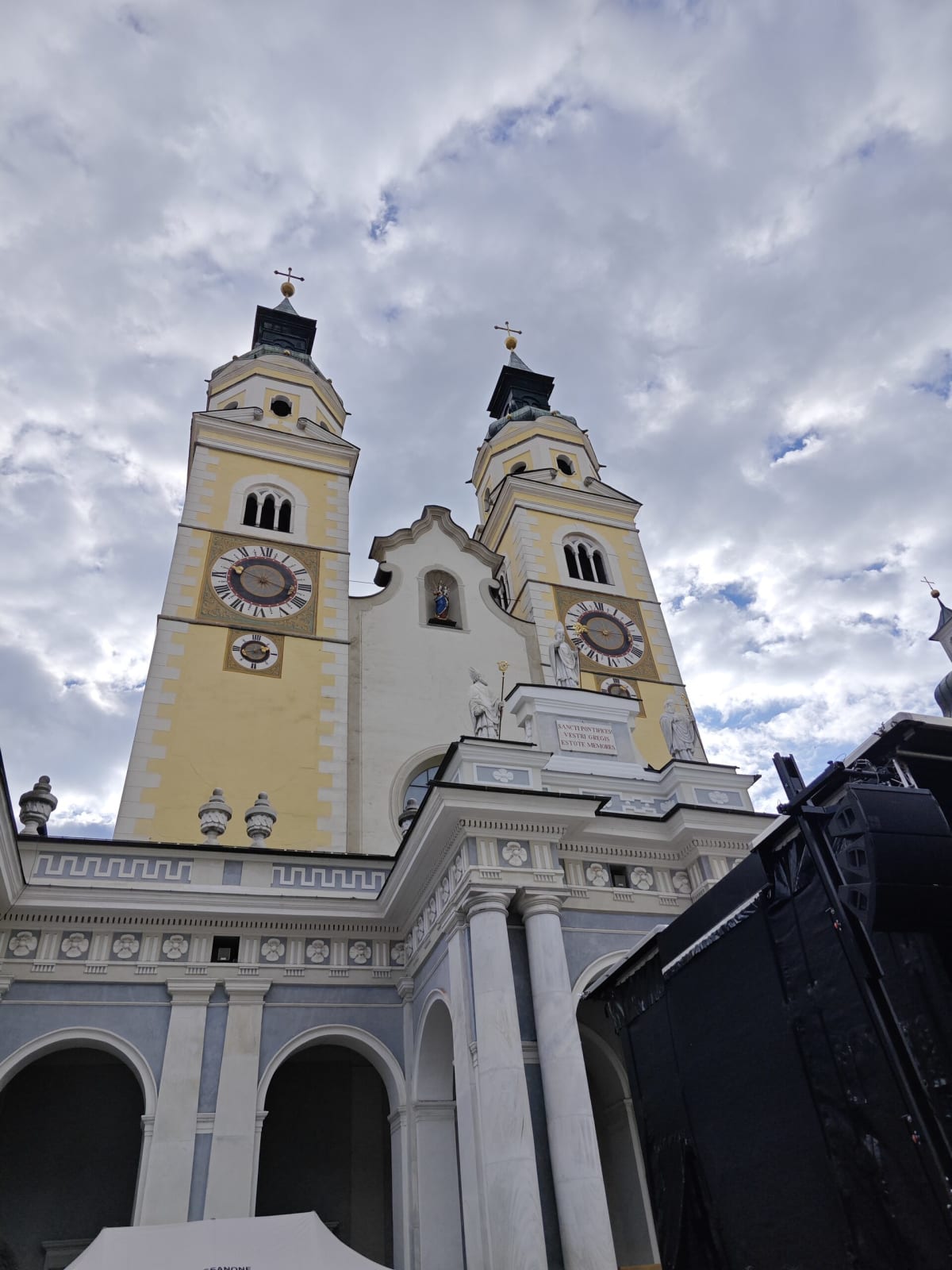 The twin yellow bell towers of Brixen Cathedral with ornate clock faces beneath a cloudy sky