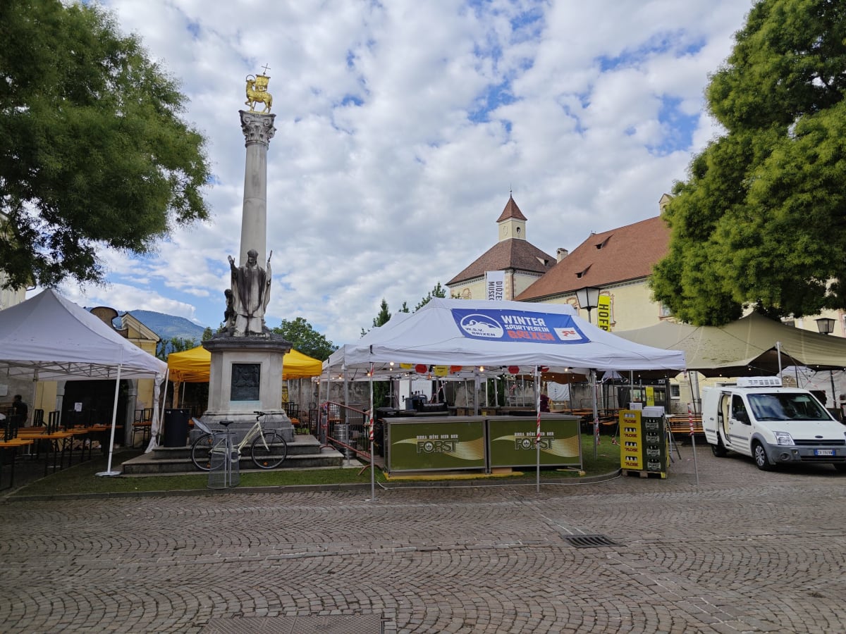 A cobbled square in Brixen with a column topped with a golden figure behind market tents under broken cloud