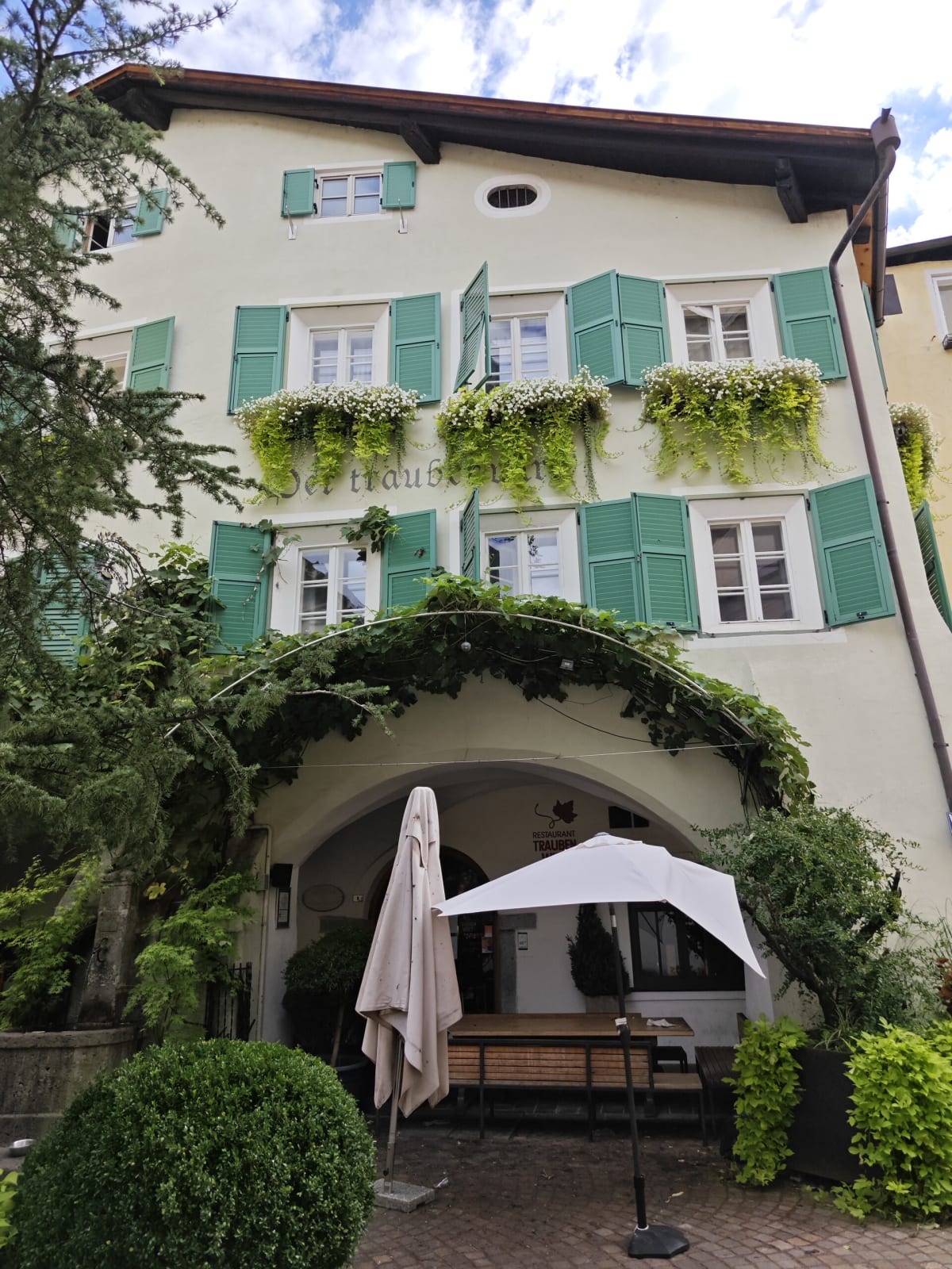 A pale townhouse with green shutters, window boxes full of flowers and a vine-covered archway over a restaurant door
