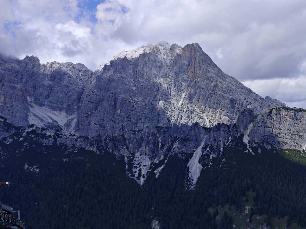 Snow-streaked grey Dolomite crags rising above shadowed pine forest, with drifting cloud