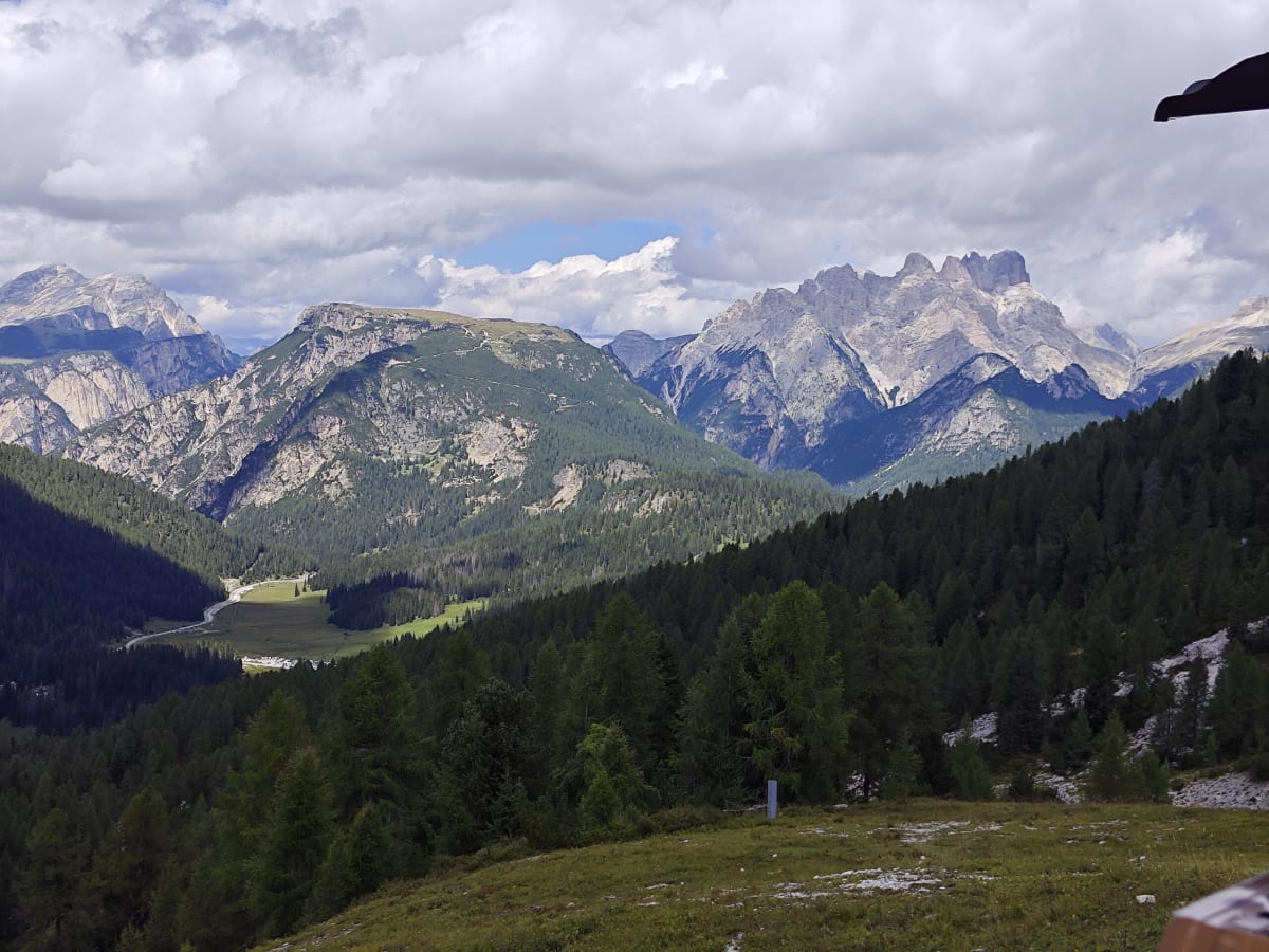 A broad panorama of Dolomite peaks across green valleys with larch woods and a mountain track in the foreground