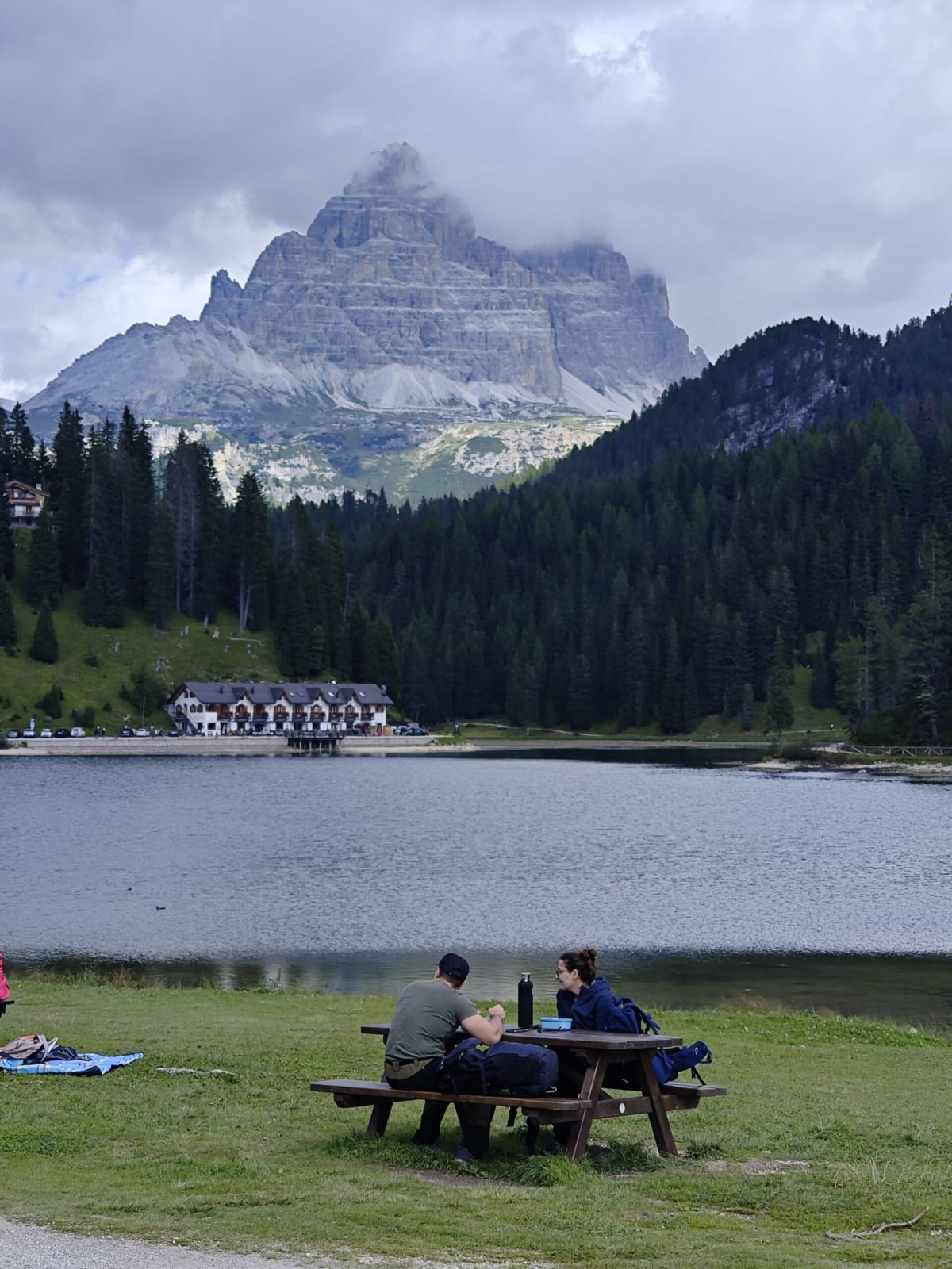 The Tre Cime di Lavaredo with cloud on their summits above Lago di Misurina, two hikers at a picnic table on the shore