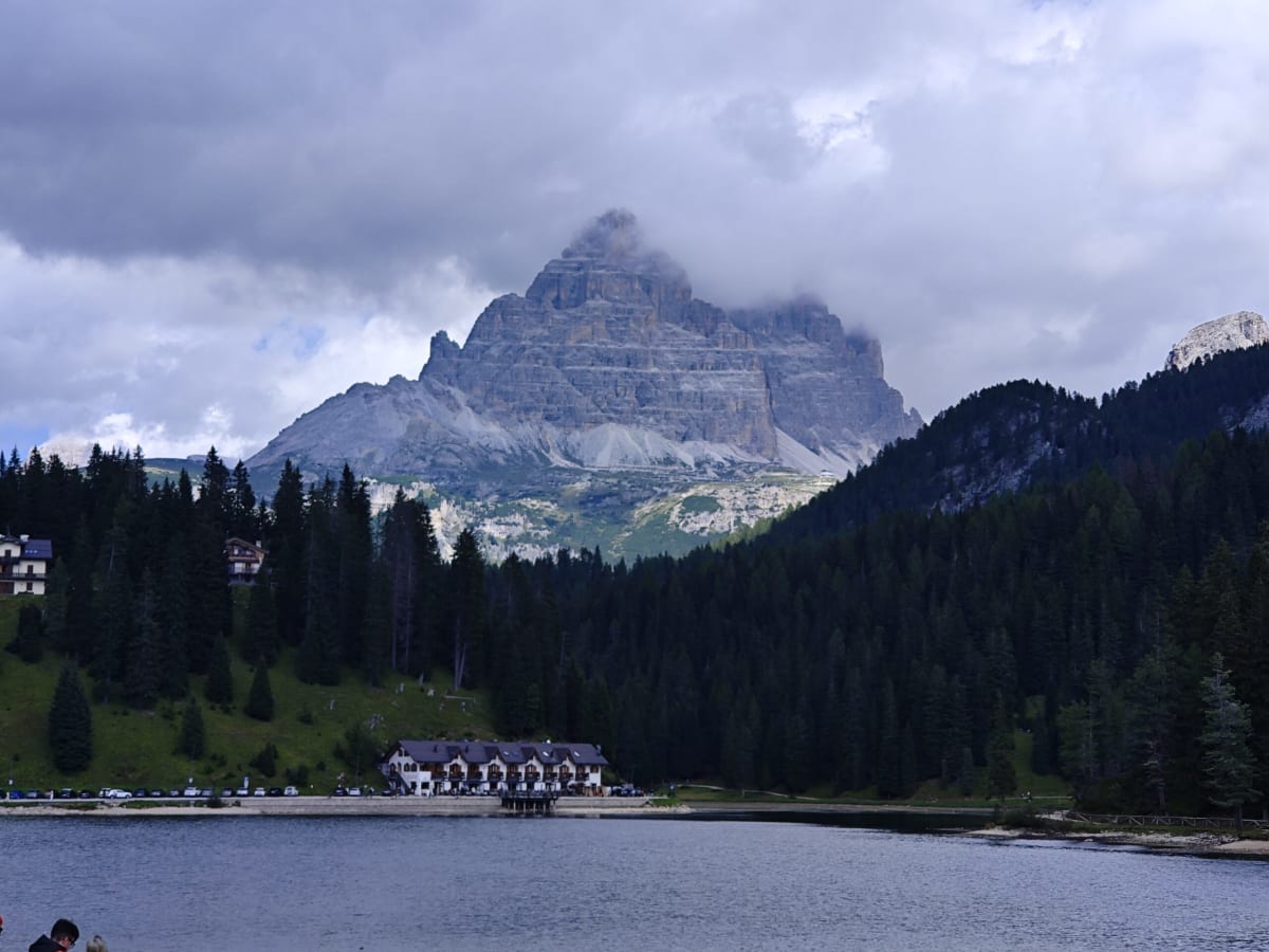The Tre Cime from across the lake, dark forest running down to a lodge at the water's edge under heavy cloud