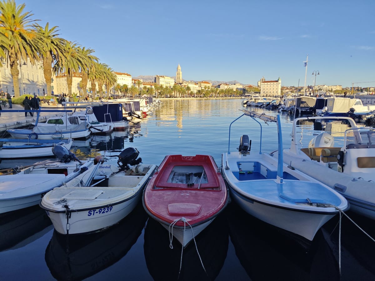 Three small open boats, white, red and blue, moored bow-on in still water, the Riva and the distant campanile framed between them