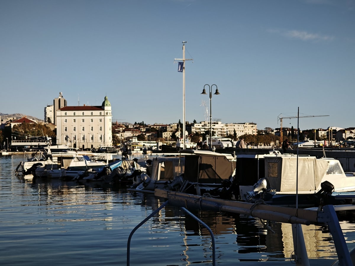 Canvas-covered boats at their moorings in late-afternoon shade, a white-domed building catching the last direct light on the far side of the harbour
