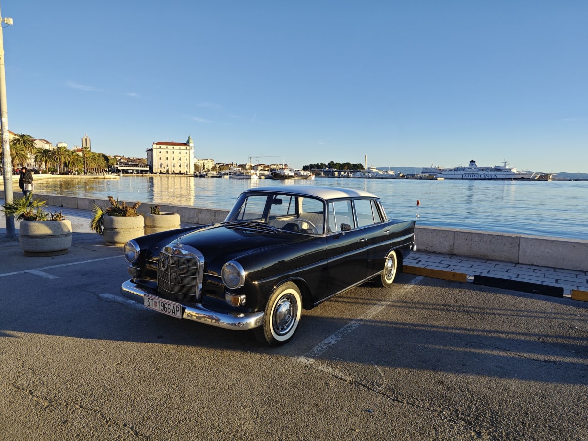 A black vintage Mercedes with a white roof and whitewall tyres parked along the Riva, the harbour and a Jadrolinija ferry behind it in flat winter light
