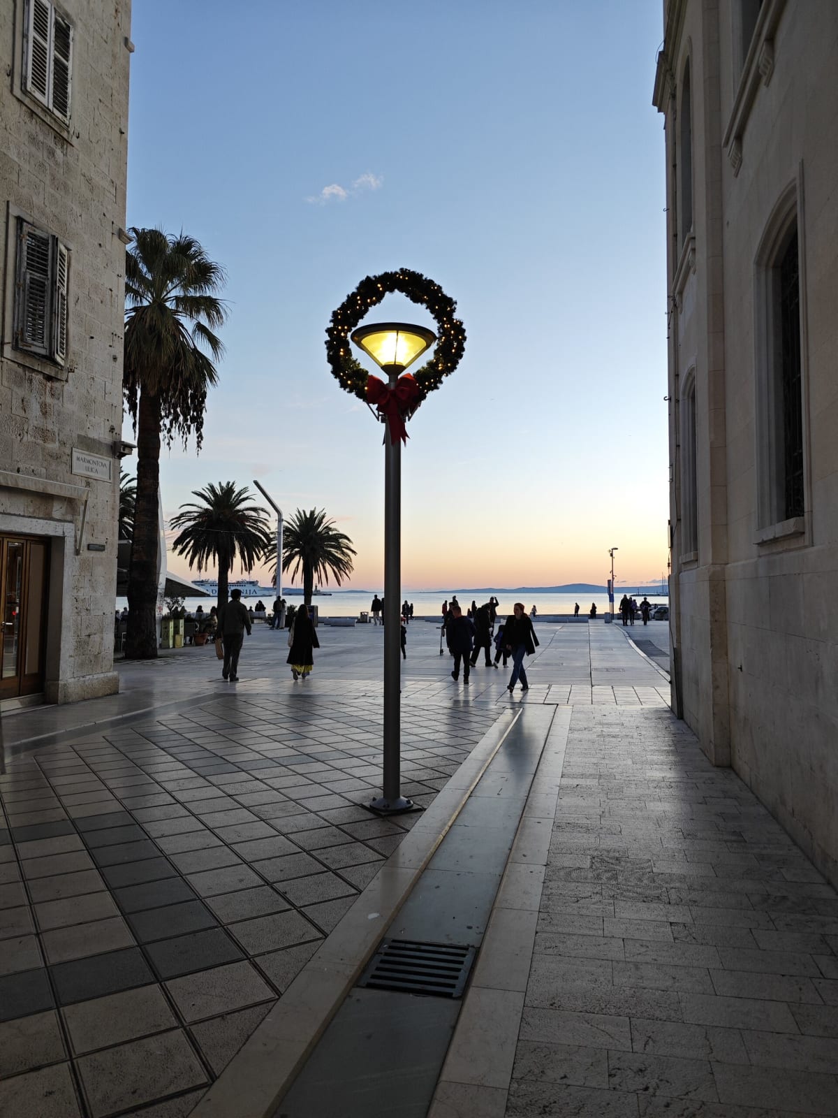 A narrow stone alley opening onto the Riva at dusk, a street lamp wrapped in a lit Advent wreath with a red bow in the centre of the frame, palms and walkers silhouetted against a pale orange sky