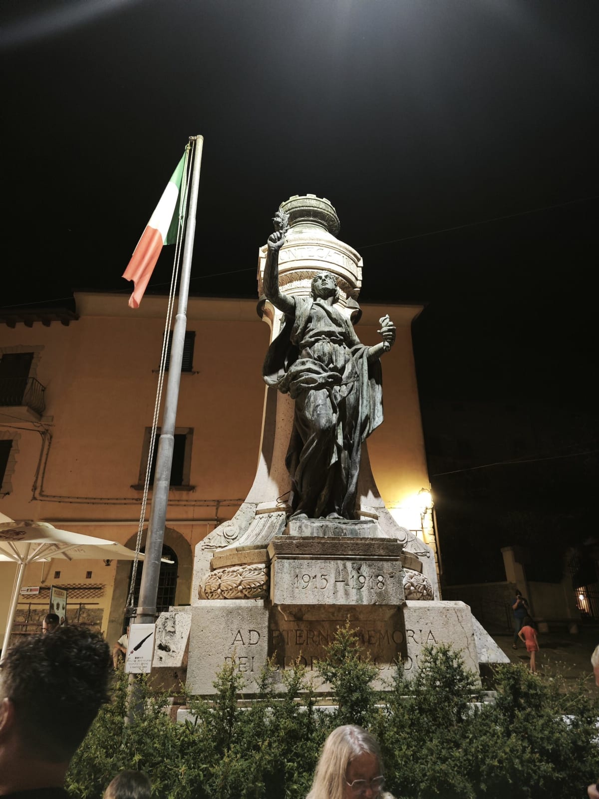 A bronze statue of a robed figure on a stone plinth, an Italian flag on a pole beside it, floodlit against the night sky