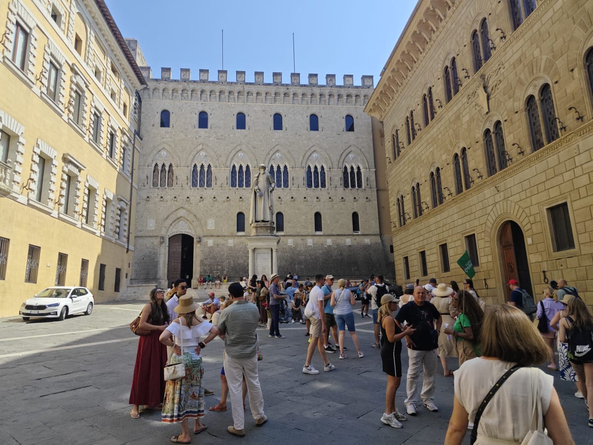 A crowded Sienese piazza in the sun, a long battlemented palazzo of pale stone at the back and ochre façades along both sides