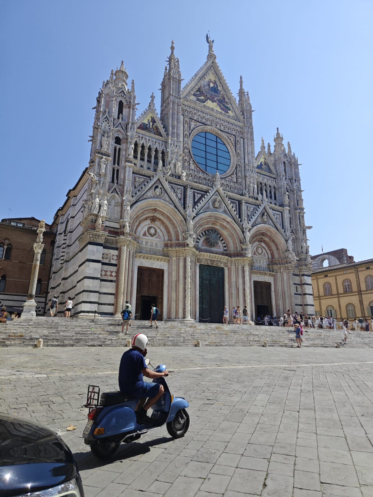 The ornate black-and-white marble façade of Siena Cathedral, a blue rose window above its three portals, a scooter crossing the stone square below