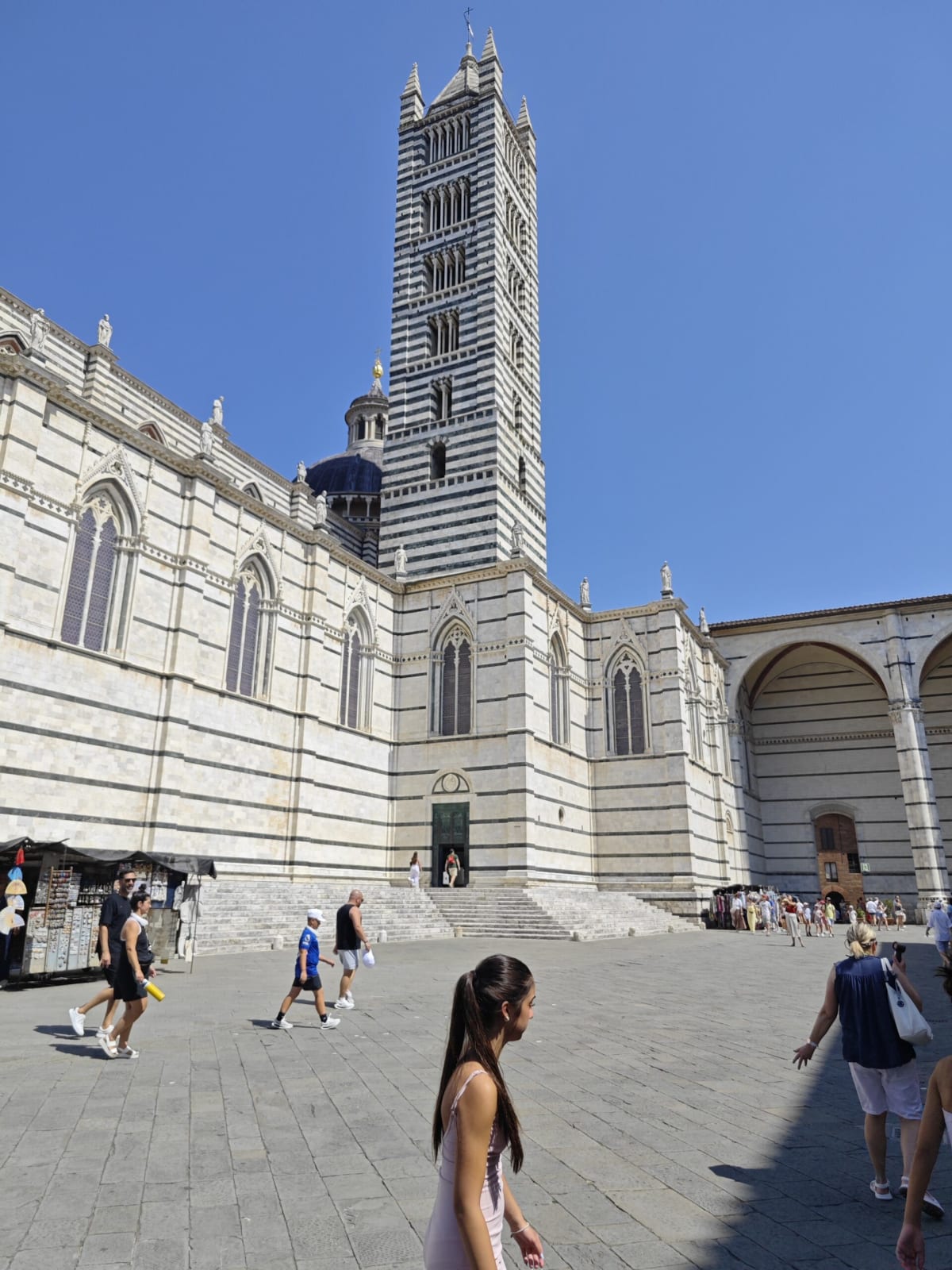 The striped marble campanile of Siena Cathedral rising behind the cathedral's long flank, tourists crossing the sunlit square