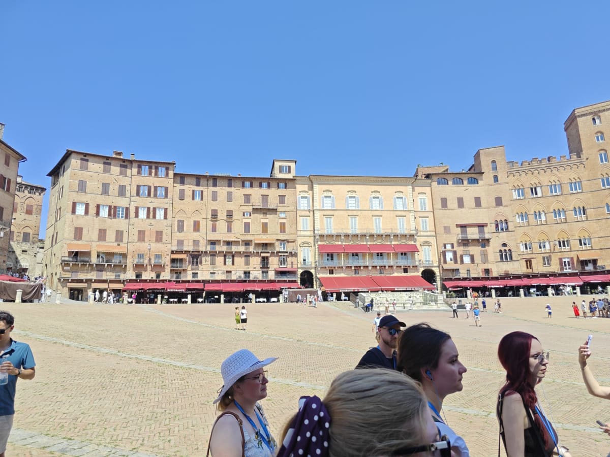 The curving row of ochre and terracotta palazzi around the Campo, café awnings along the base and visitors in the foreground