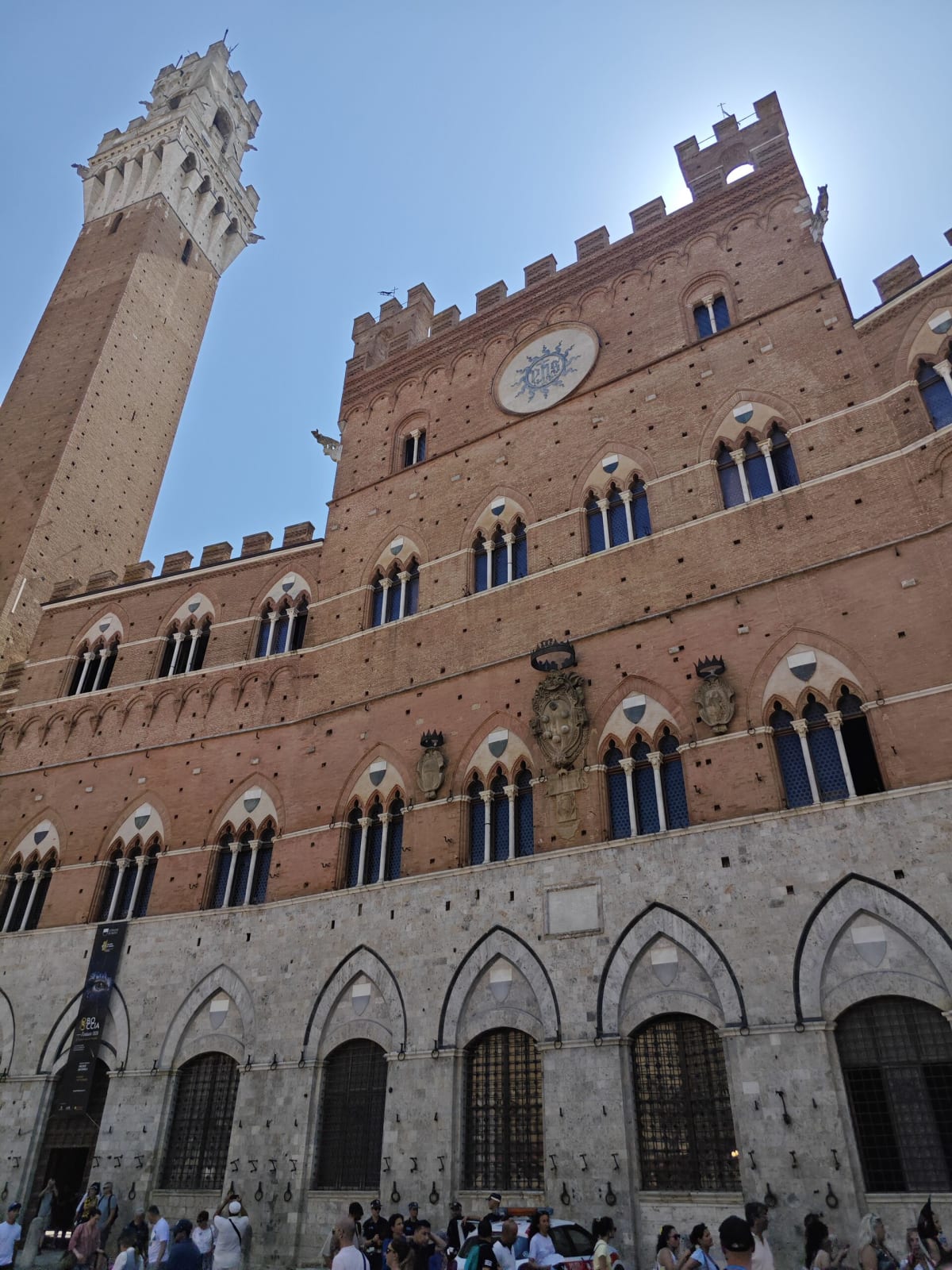 The Palazzo Pubblico from beneath, its brick façade of pointed Gothic windows and battlements, the Torre del Mangia soaring on the left