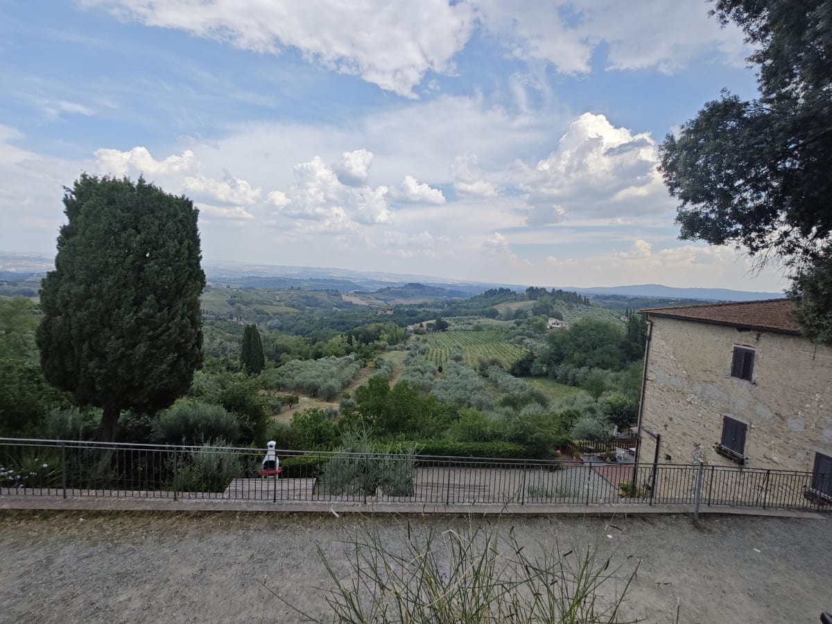 A Tuscan hillside viewpoint from a roadside terrace — cypresses, olive groves and wooded ridges rolling off beneath scattered clouds