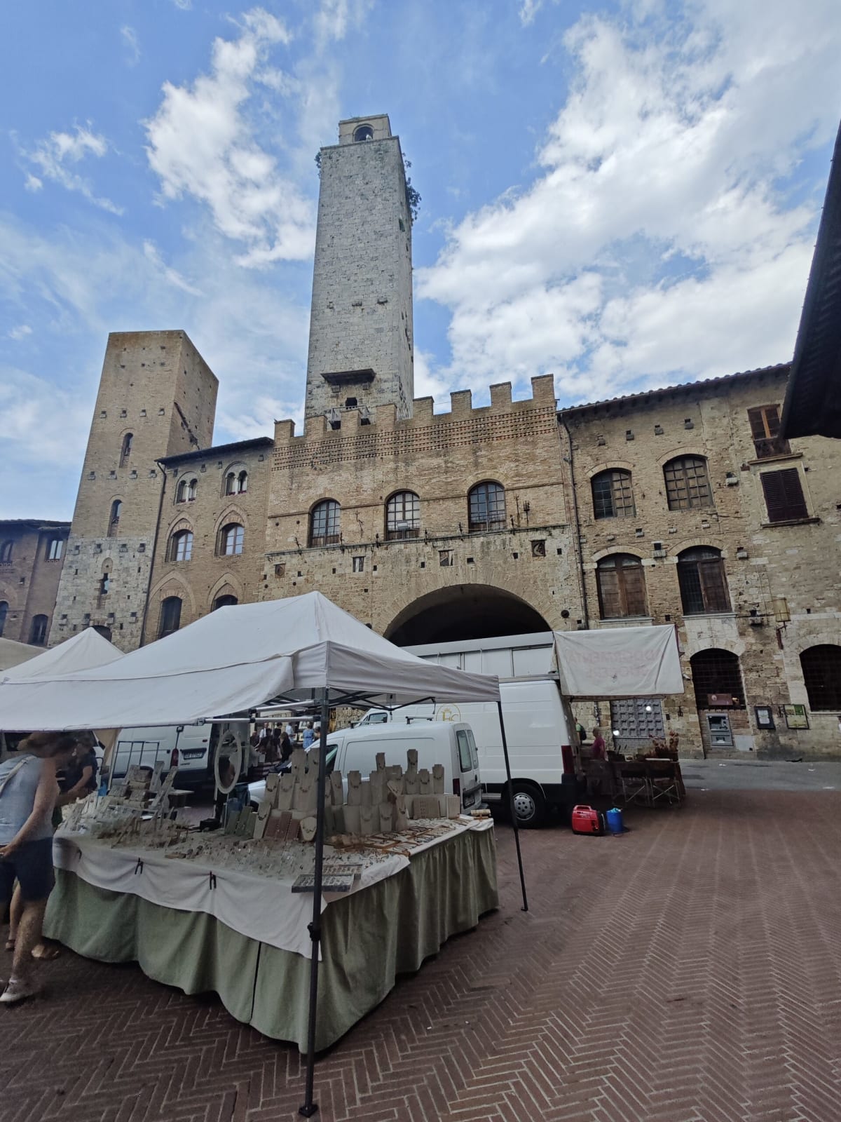 A town square in San Gimignano with a stone tower and crenellated palazzo behind a market stall under a partly cloudy sky
