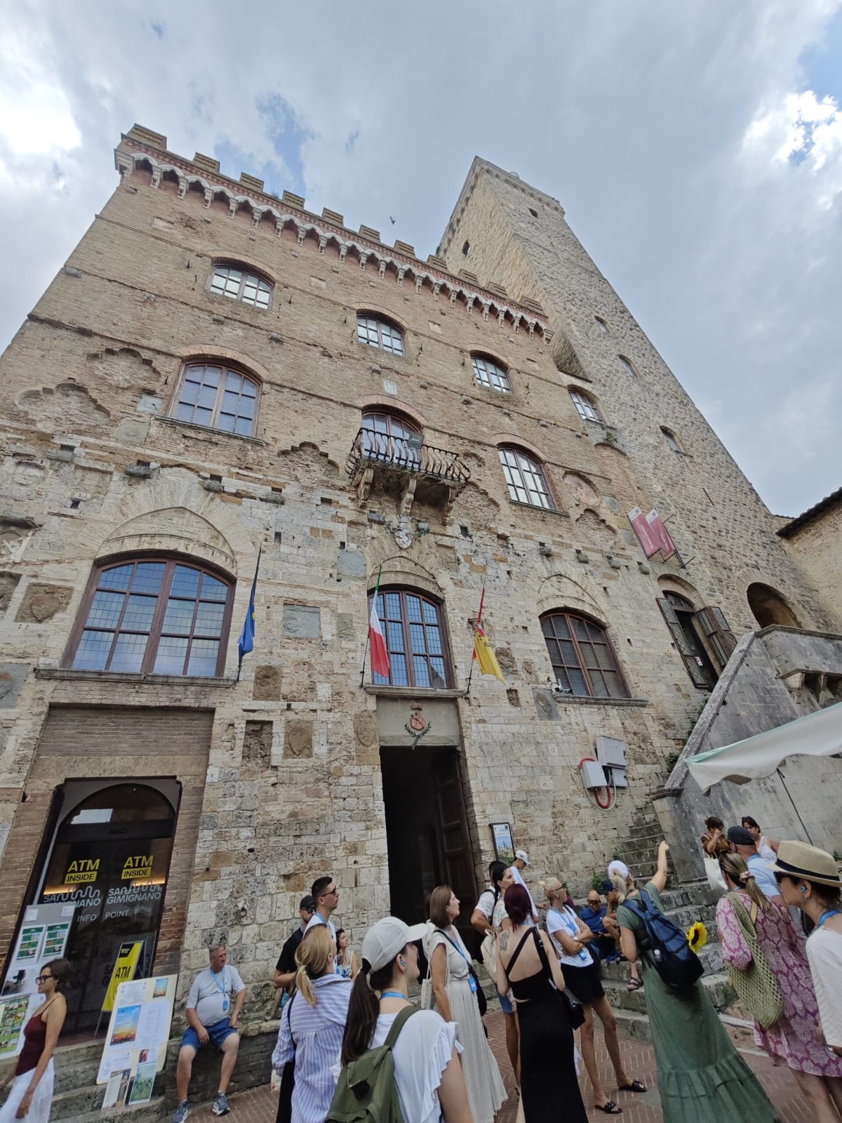 Looking up at the heavy medieval stone façade of a San Gimignano palazzo with a battlemented top and a tower behind, a tour group at the door