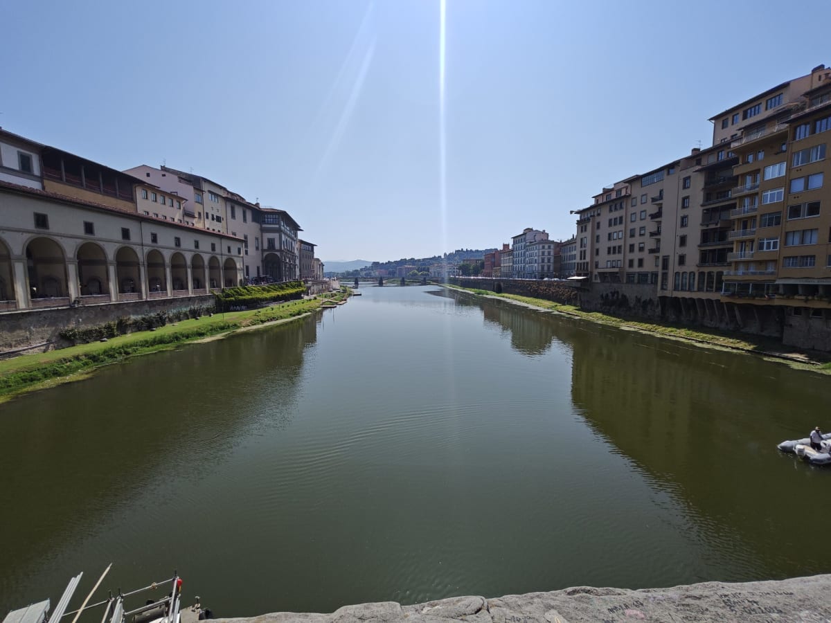 The River Arno in Florence from a bridge, calm green water between stone embankments and a loggia at the left, sun flaring over the water