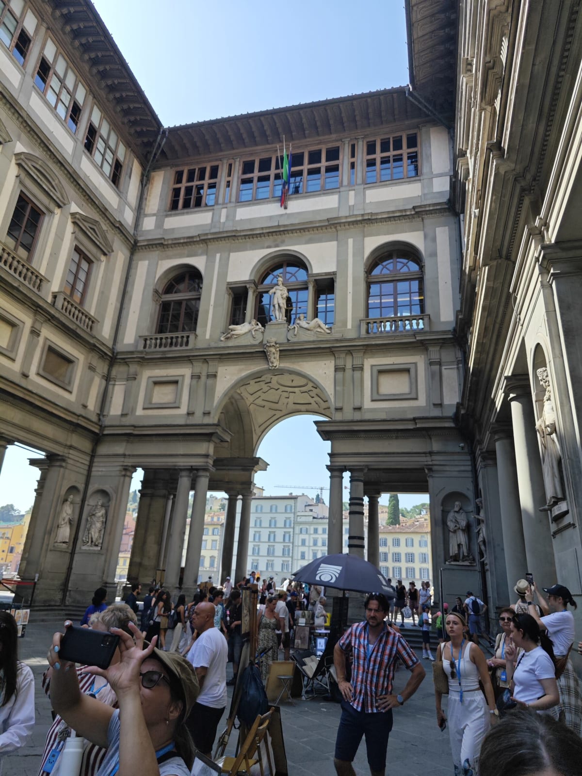 The courtyard of the Uffizi, a long arcaded passage with statues in niches and a crowd gathered at the arch opening toward the river