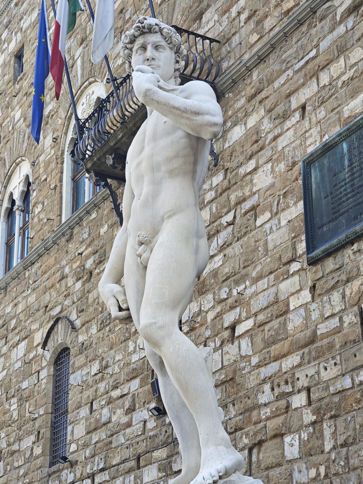 A white marble copy of Michelangelo's David raised above the street before Palazzo Vecchio, flags hanging from the façade behind
