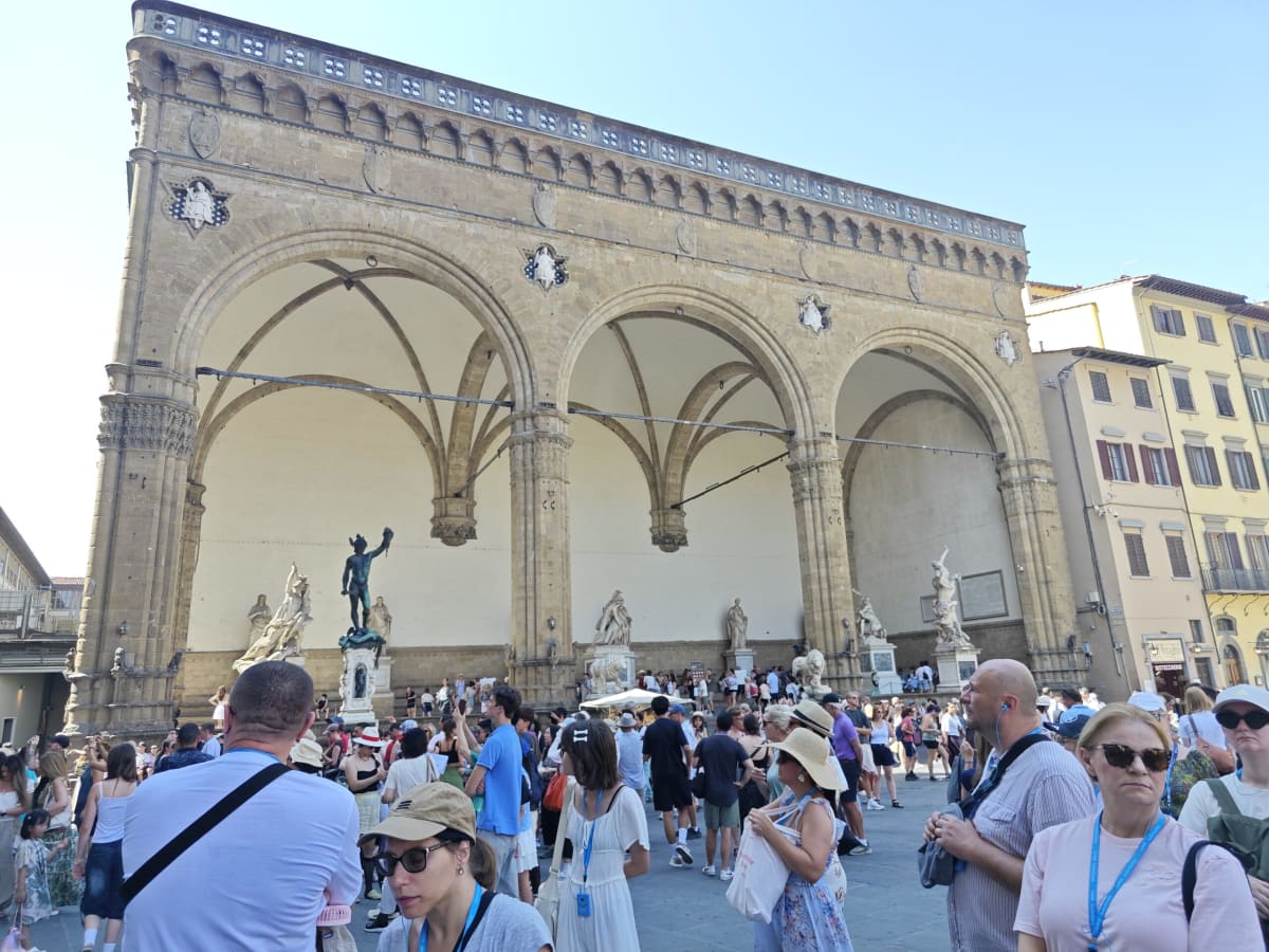 The Loggia dei Lanzi, a great arcade with three arches filled with sculpture, and a crowd of tourists in the sunlit Piazza della Signoria
