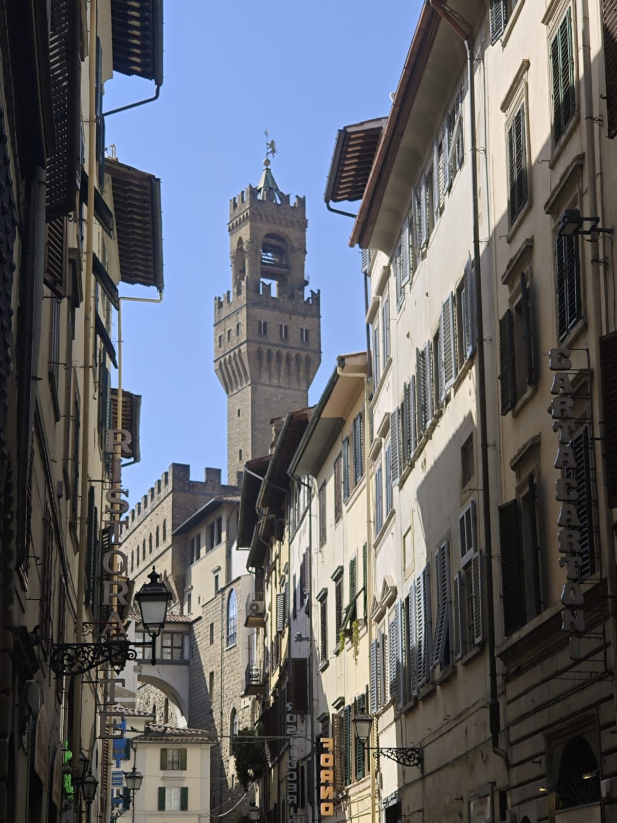 The tower of Palazzo Vecchio rising above a narrow Florentine street, balconies and shutters along both sides
