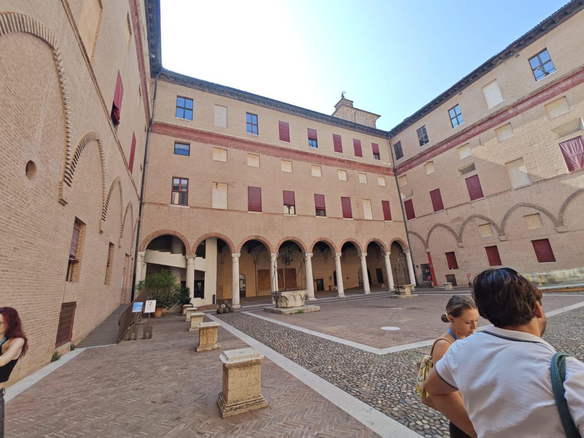 An inner courtyard of Ferrara's Castello Estense, an arcaded brick loggia around a cobbled square and a few visitors