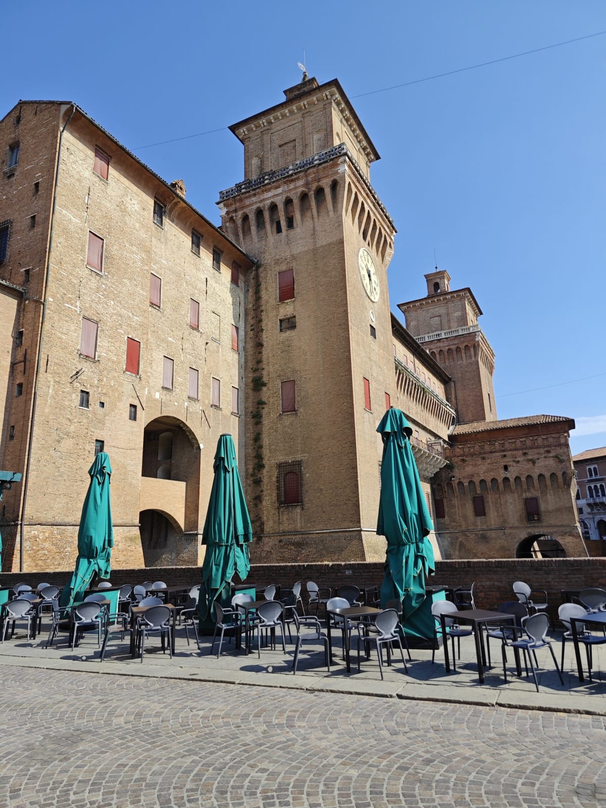 A massive brick tower of the Castello Estense beside café tables with folded green umbrellas on the cobbles