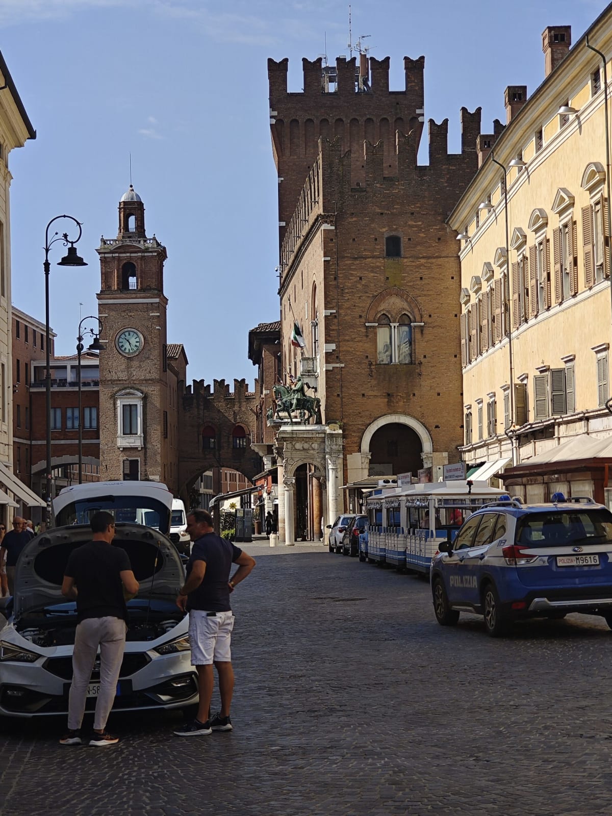 A street in Ferrara with a clock tower and a battlemented brick palazzo behind a couple of police cars