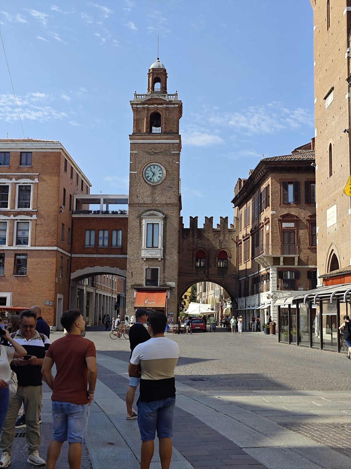 A slim brick clock tower rising over a gateway arch between old Ferrara houses, pedestrians in the foreground