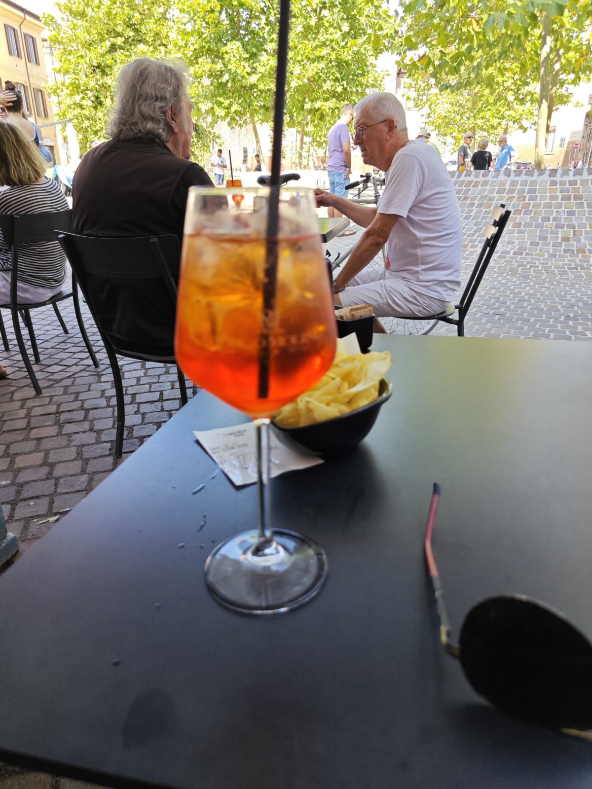 An orange Aperol spritz with a bowl of crisps on a café table, two men sitting talking at the next table on the cobbled square