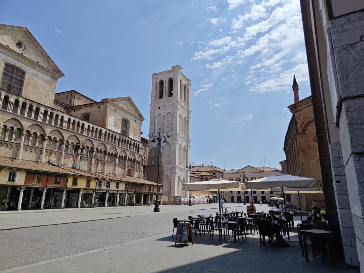 Ferrara's cathedral square with the white campanile behind a long arcaded row of shops and café tables in the sun