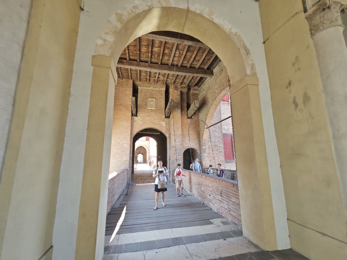 A covered brick passage with a timber ceiling leading into a castle courtyard, visitors walking through