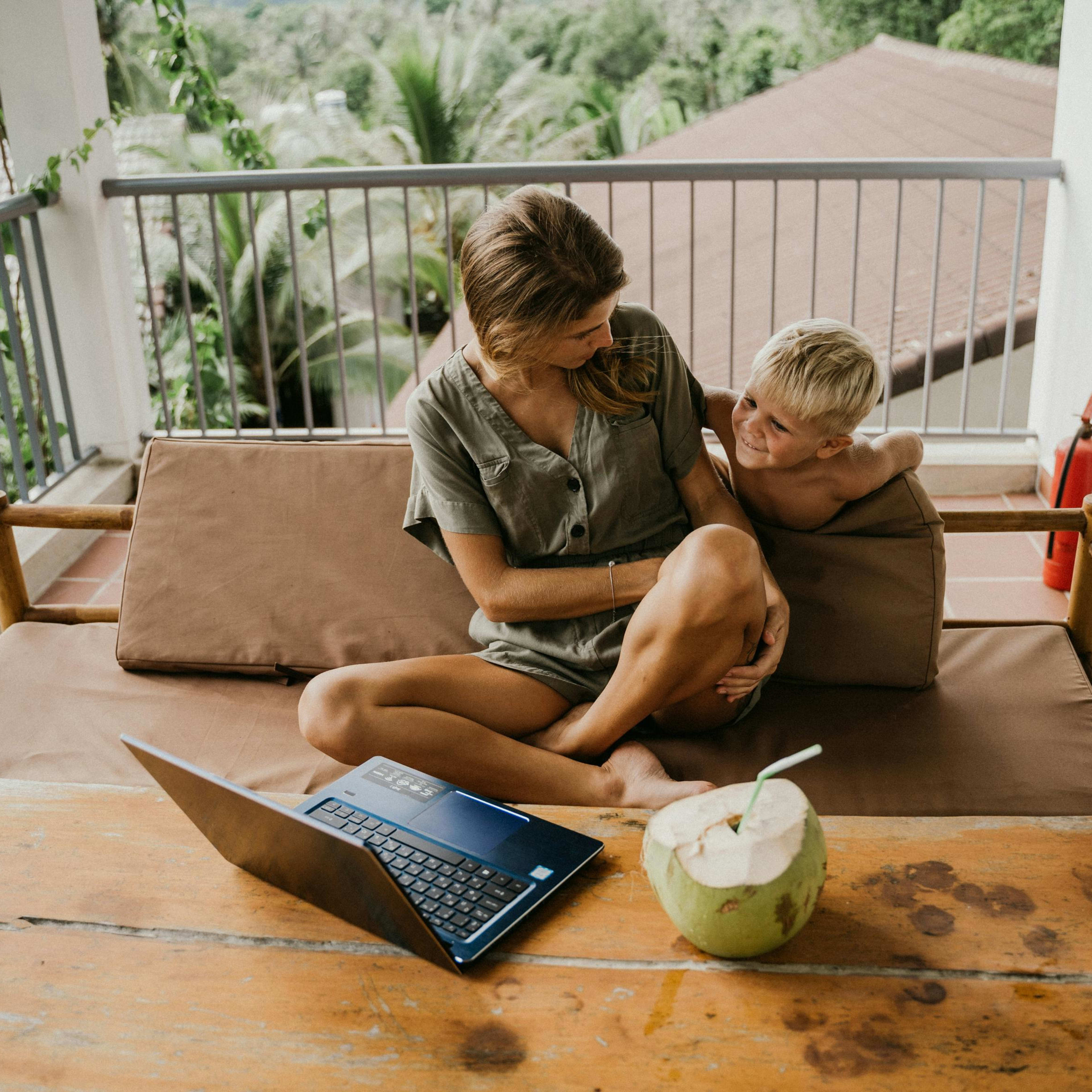 A woman and a young boy sit on a lounge chair on a balcony, with a laptop and a coconut on a wooden table in the foreground and lush greenery and rooftops in the background.