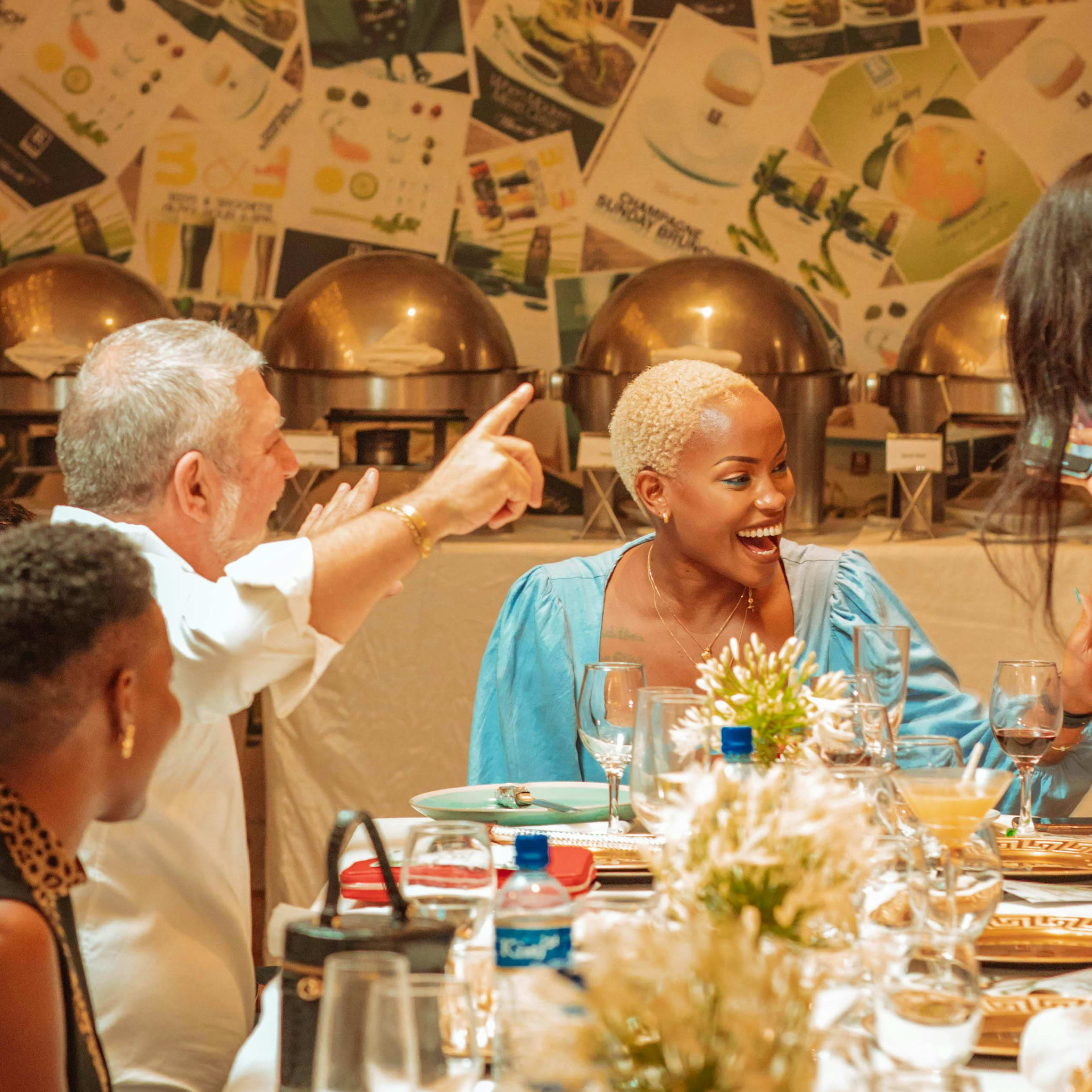 A man points toward a laughing woman seated at a dinner table set with glassware and flowers, all positioned in front of a wall covered in posters and buffet warming trays.