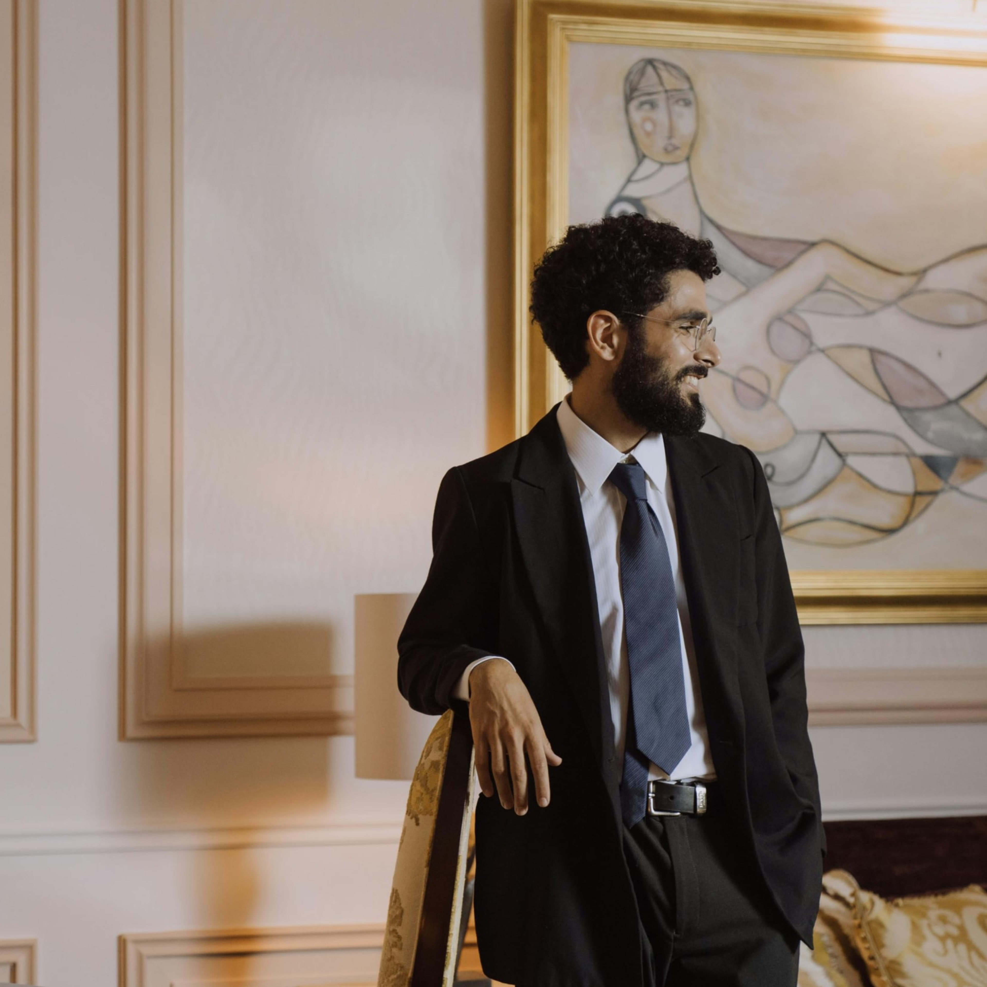 A bearded man in a black suit and blue tie leans against a chair in a room featuring a large painting and ornate wall molding.