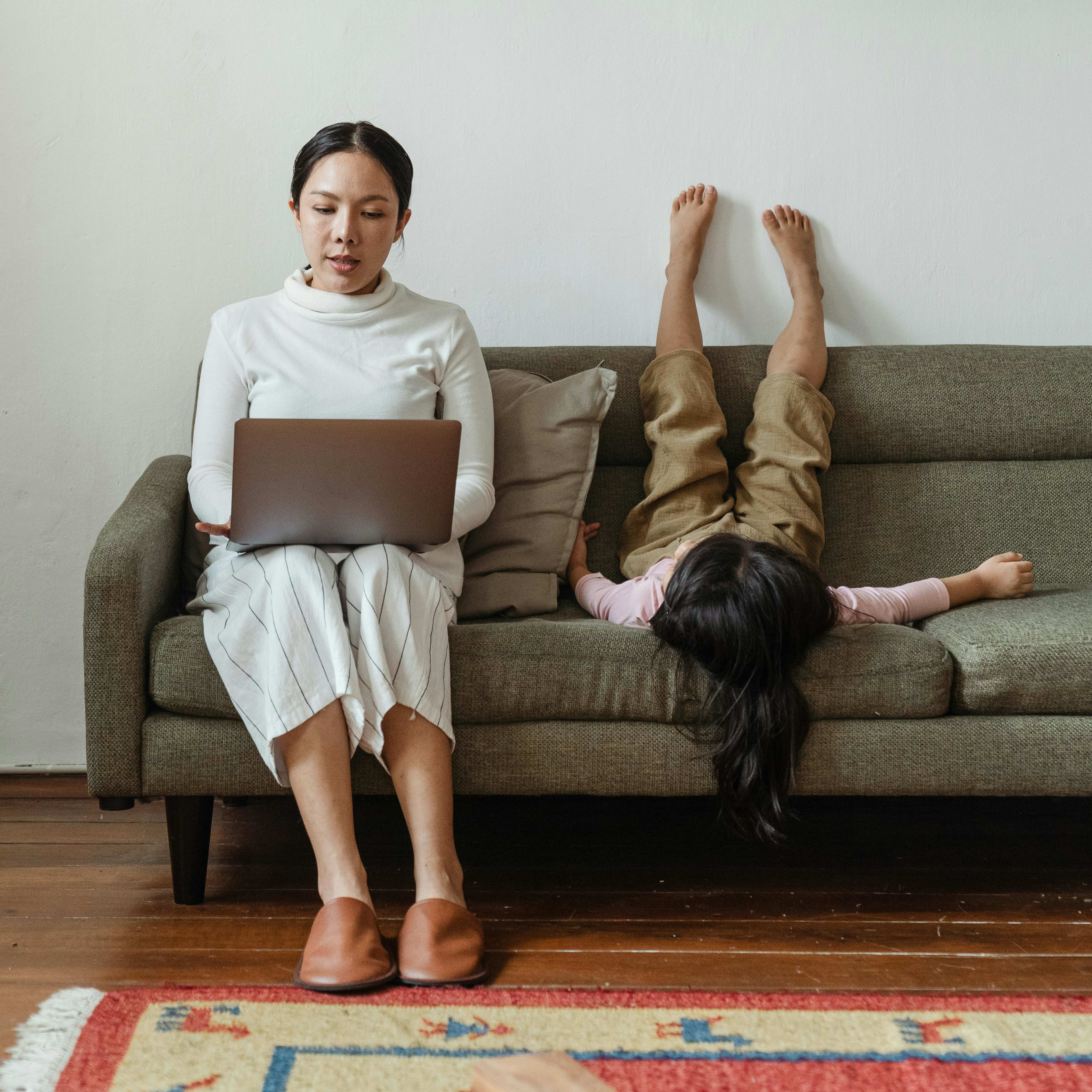 A woman works on a laptop while a child rests upside down on a green sofa against a plain white wall.