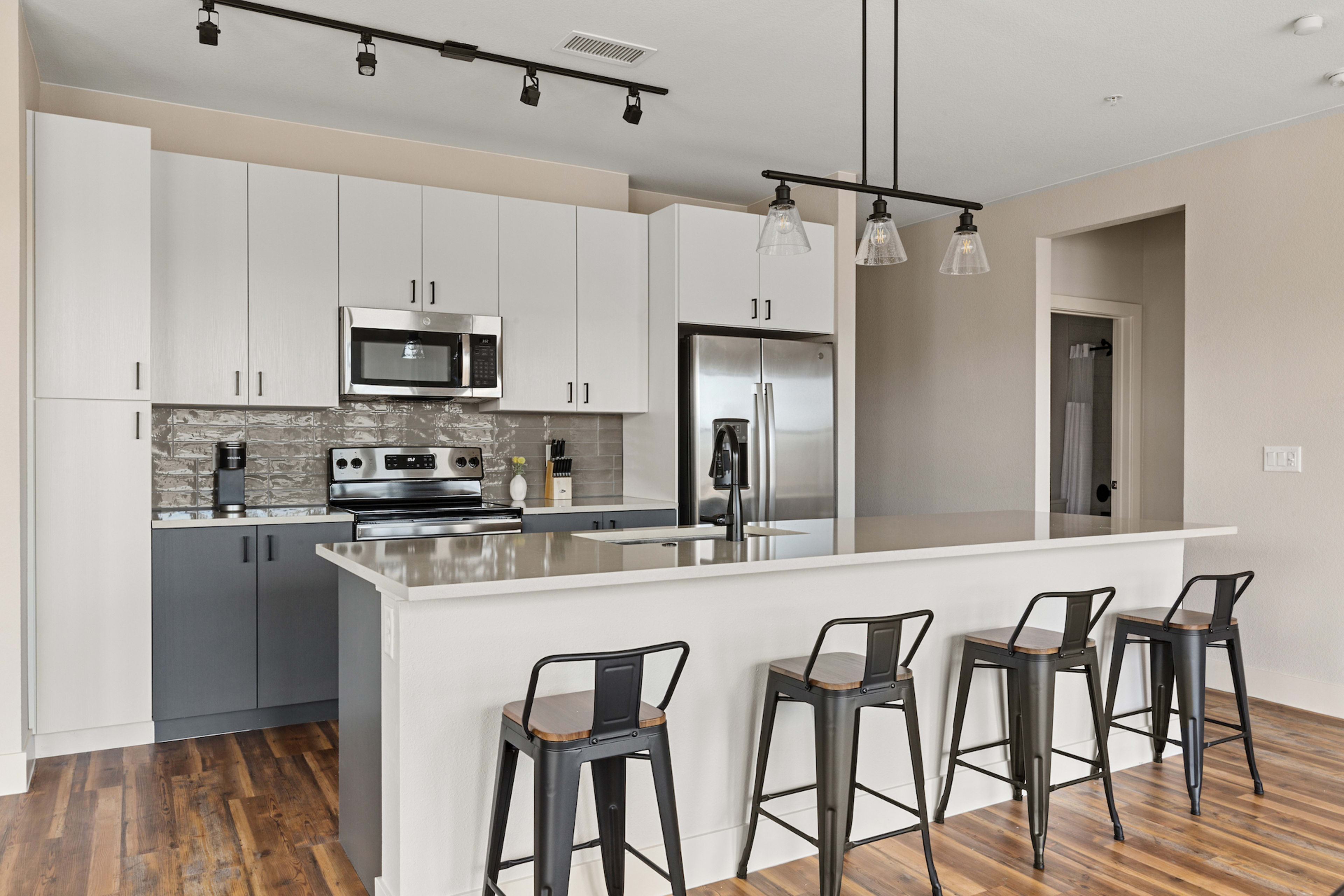 Four black barstools sit before a white kitchen island in the foreground, with white and grey cabinetry and stainless steel appliances in the background.