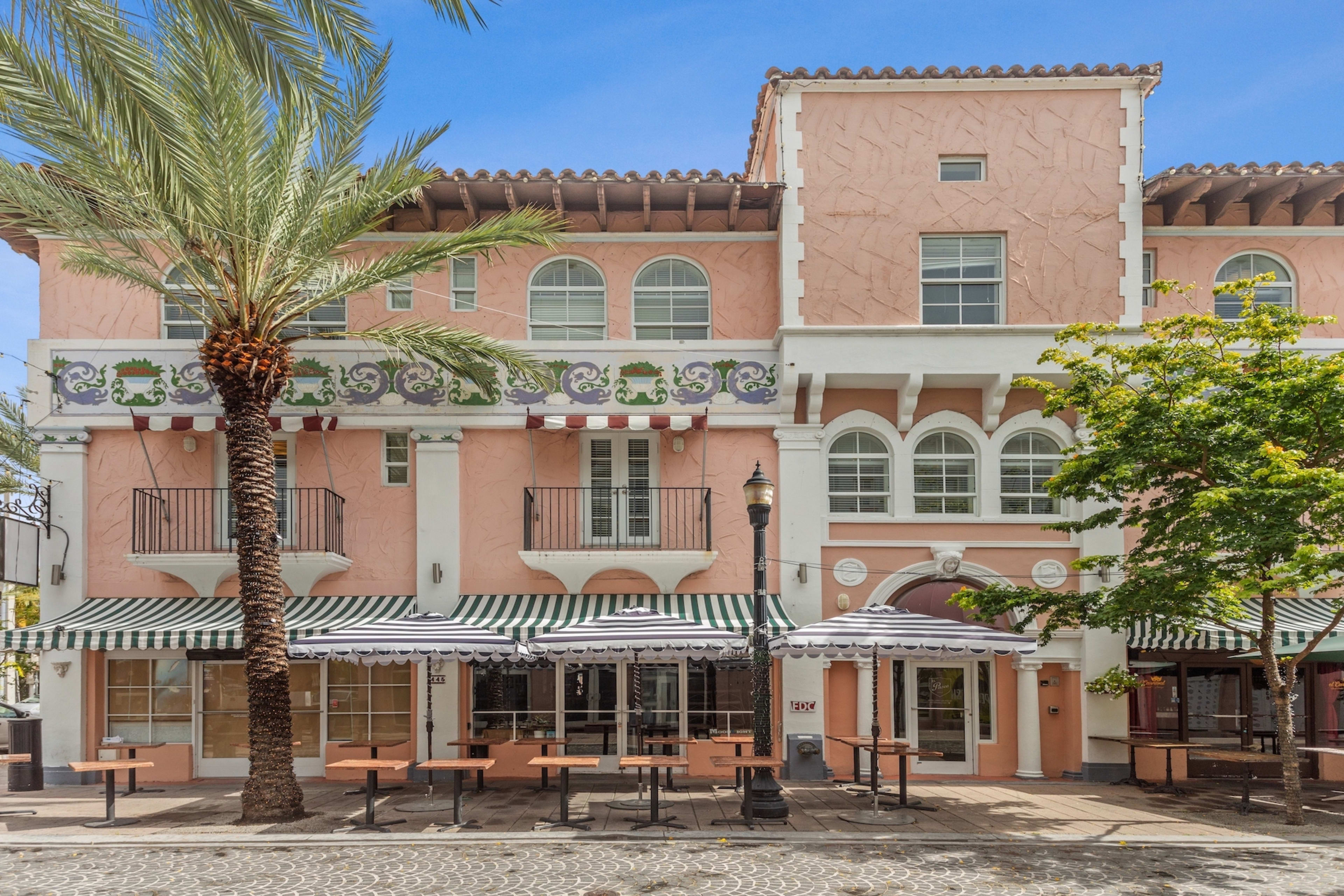 An outdoor dining area with tables and umbrellas sits in the foreground before a pink, Mediterranean-style building under a clear blue sky.