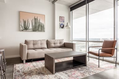 A modern living room features a beige sofa, a dark coffee table, and a brown leather chair on a patterned rug, with a large window overlooking a city skyline in the background.