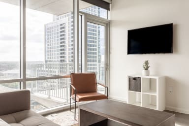 A modern living room featuring a brown leather chair and grey coffee table in the foreground, with a wall-mounted TV and a view of city skyscrapers through large windows in the background.