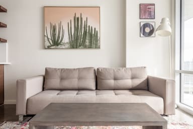 A grey concrete coffee table sits in the foreground before a beige tufted sofa, with a cactus painting and small artworks adorning the white wall in the background.