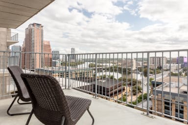 Two brown wicker chairs sit on a concrete balcony overlooking a city skyline and a river under a cloudy sky.