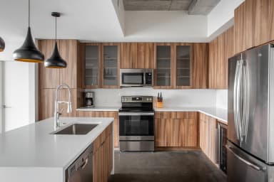 A modern kitchen features a white island with a stainless steel sink in the foreground and wood cabinetry with stainless steel appliances in the background.