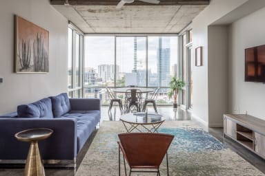 A blue velvet sofa and leather chair sit on a patterned rug in the foreground of a modern living room, with a dining set and floor-to-ceiling windows overlooking a city skyline in the background.